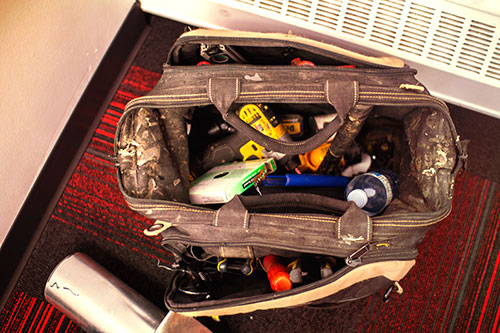 A worn black tool bag filled with various tools and a water bottle sits on a red and black patterned carpet next to a metal tumbler, ready for another day with Family Plumbing and Heating.