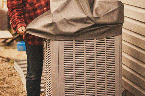 A person places a cover over an outdoor air conditioning unit next to a house, ensuring protection and efficiency—just one of the quality services offered by Family Plumbing and Heating.
