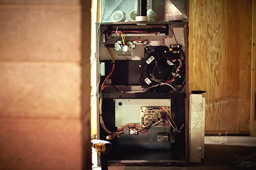 Open furnace unit in a basement, showing internal components, wiring, and circuit boards, with unfinished wooden walls and a concrete pillar nearby—serviced by Family Plumbing and Heating.