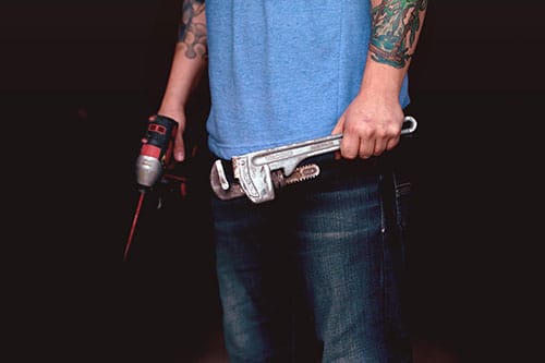 A person wearing a blue shirt and jeans holds a pipe wrench and a power drill, tattoos visible on both arms, ready for any Family Plumbing and Heating job.