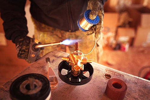 A technician from Family Plumbing and Heating solders a copper pipe fitting with a torch and soldering wire on a workbench; tools and materials are visible nearby.