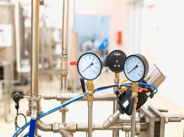 Two pressure gauges are mounted on industrial pipes, displaying readings. Stainless steel pipes and valves are visible in a clean, brightly lit facility—a testament to the quality of Regina plumbing services.