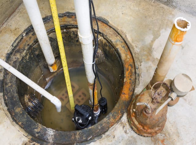 A sump pump system installed in a concrete basement floor, with various pipes and standing water visible inside the pit—expertly maintained by Regina plumbing services.