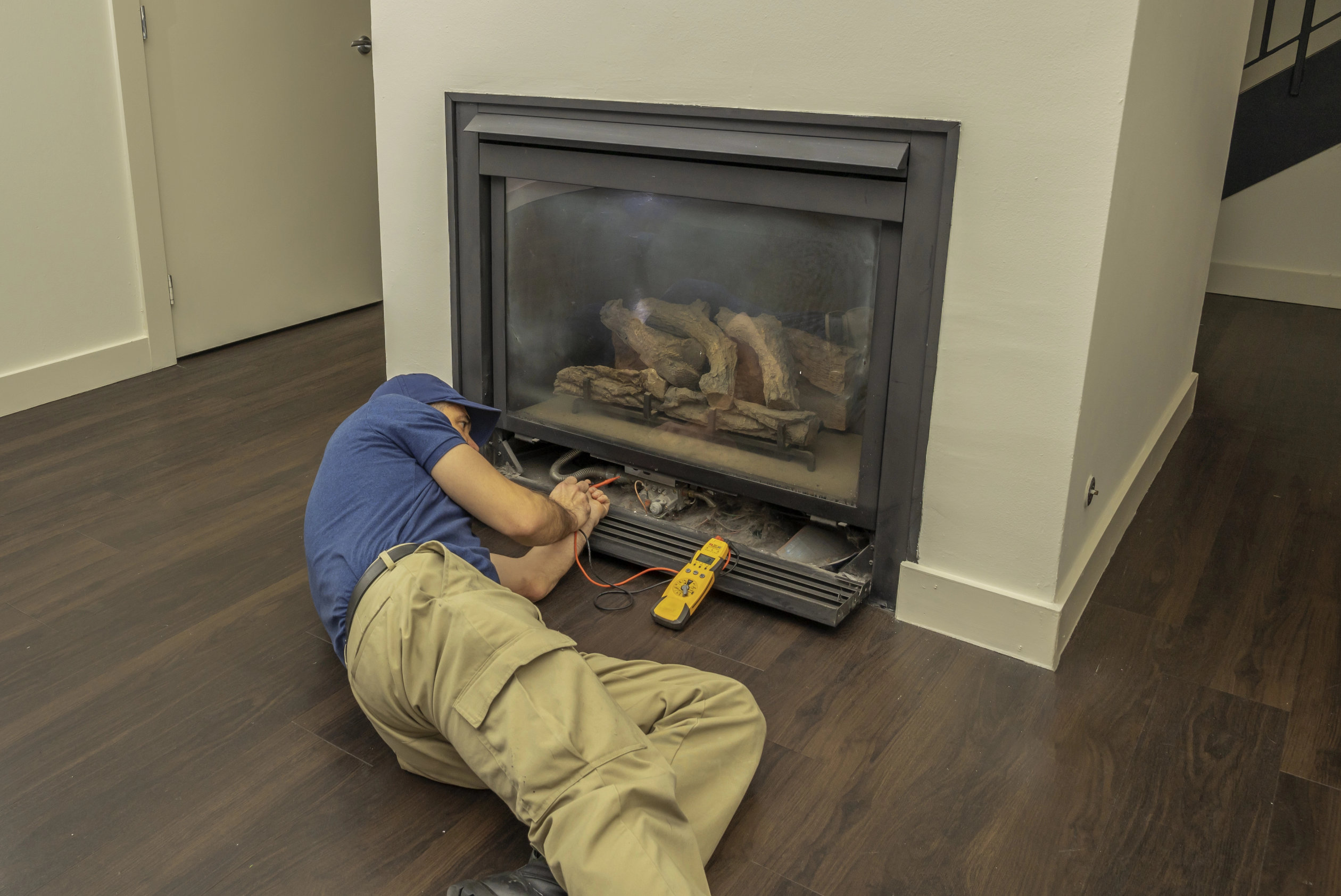 A person in a blue shirt and khaki pants uses a multimeter to inspect or perform gas fireplace repair in a modern room with dark wood flooring.